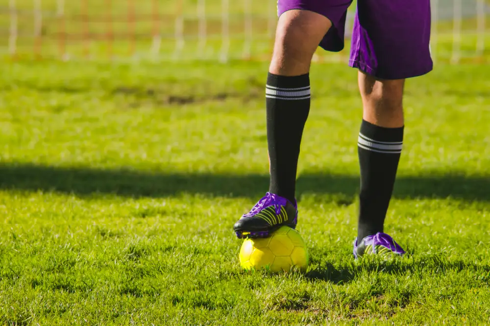 picture of a man with 1 foot on a football standing on grass wearing a purple football kit