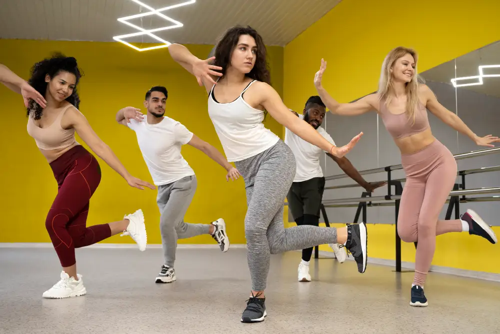 3 women and 2 men dancing in a group in a dance studio with yellow walls and and a mirror on 1 wall