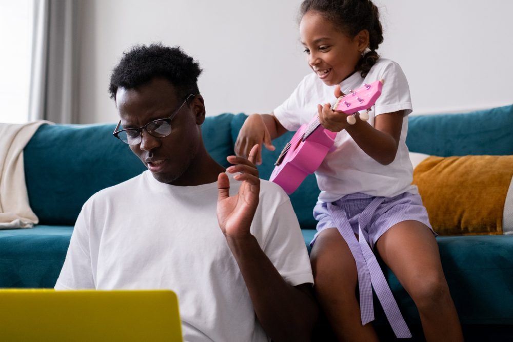 A man sitting on the floor. He is looking at his laptop and concentrating. Behind him on the sofa his daughter trying to play toy guitar
