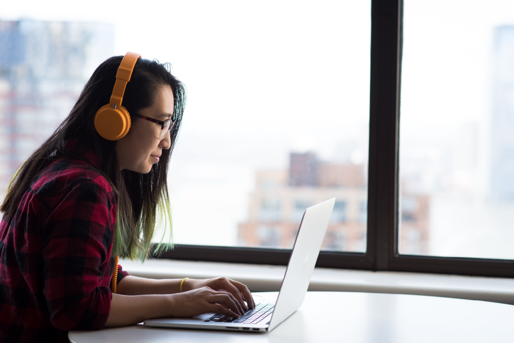 A girl in orange headphones is sitting at the table in front of her laptop. Behind her there is a big window