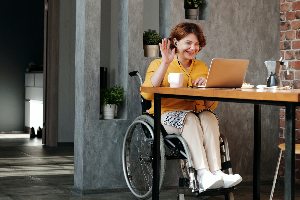 A girl in a yellow shirt in a wheelchair is waving at her laptop screen. She is wearing headphones.