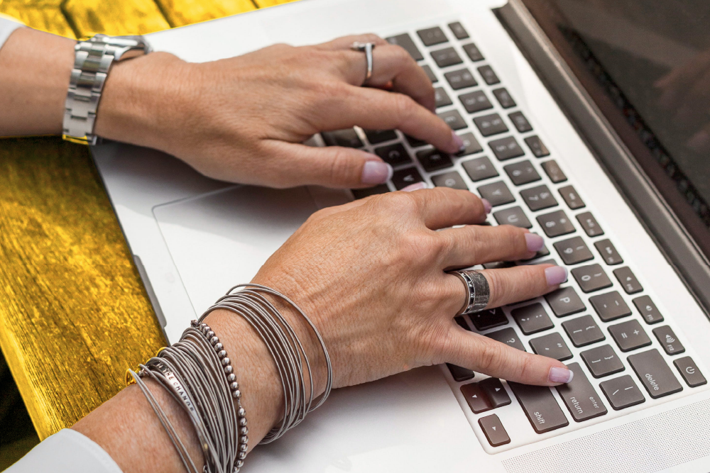 A woman who is wearing lots of metallic bracelets on one hand and watch on the other is typing on a laptop keyboard. 