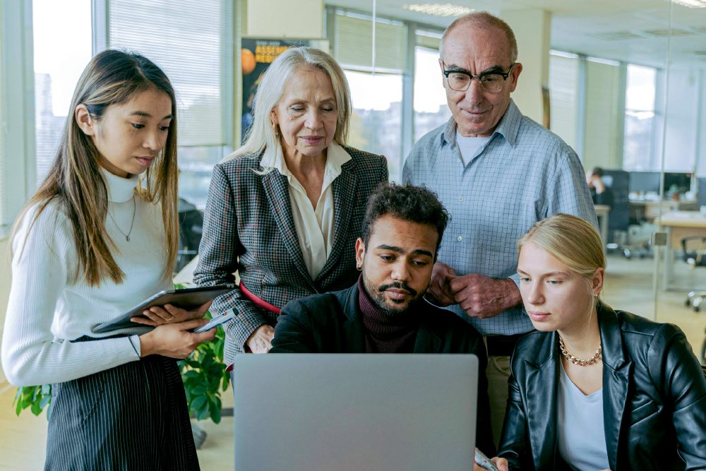 there is a man sitting at the desk and looking at the laptop. A woman is sitting next to him. Older man and older woman are standing behind him and looking at his laptop screen