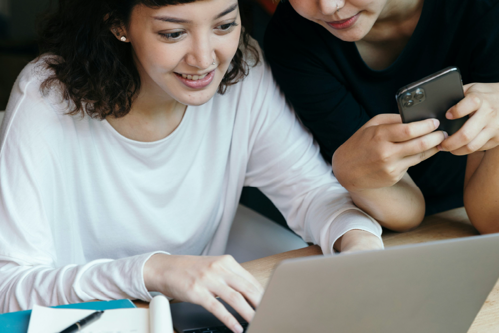 A girl in white top is sitting at the desk and looking at the rlaptop. She is smiling. There is a girl in a black top standing next to her 
