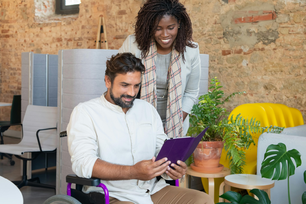 A man in a white shirt is sitting in a chair. He is holding a tablet in a purple cover. A woman in a grey sweater is standing next to him