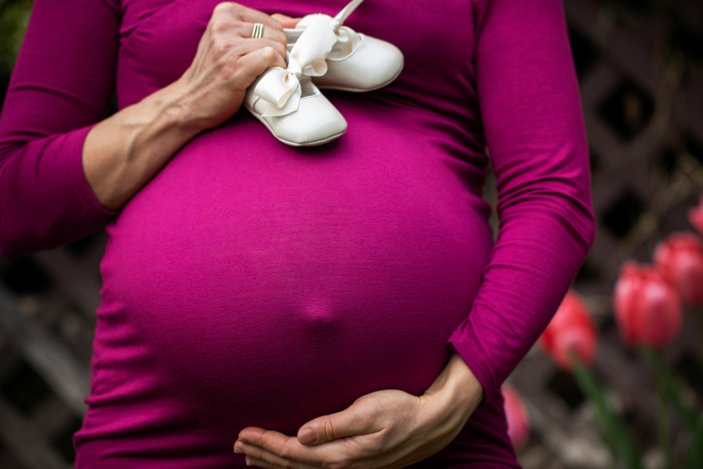 A woman wearing a purple top holding her pregnant belly and holding a pair of white baby shoes