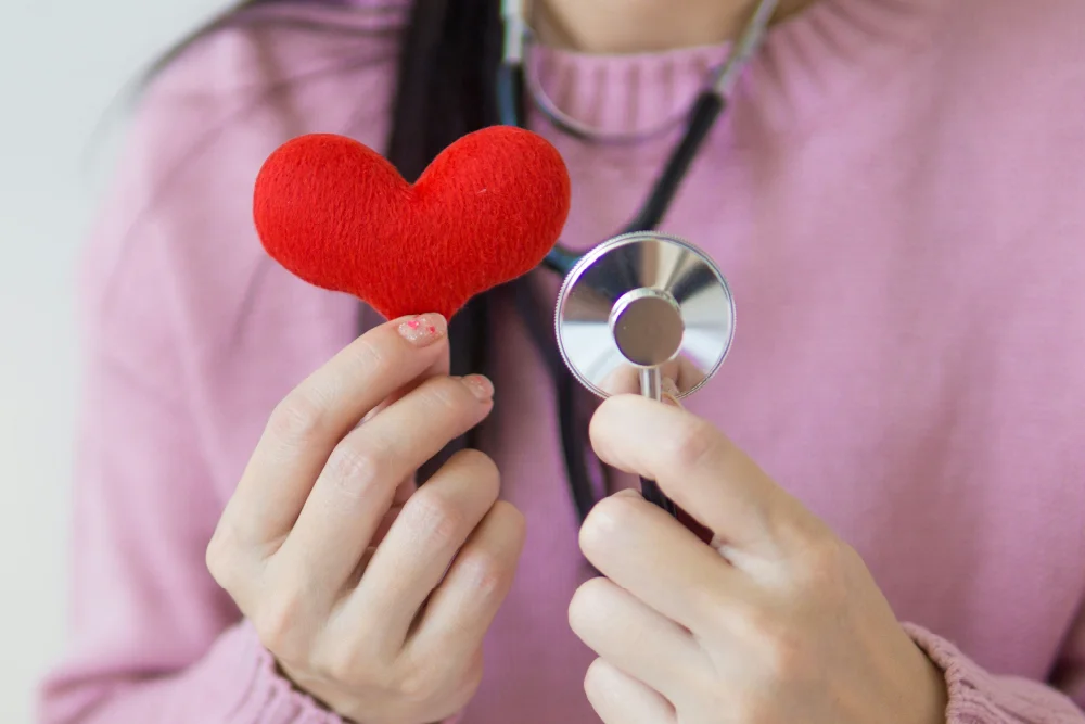 A woman in a light pink sweater is holding a red toy heart and a stethoscope
