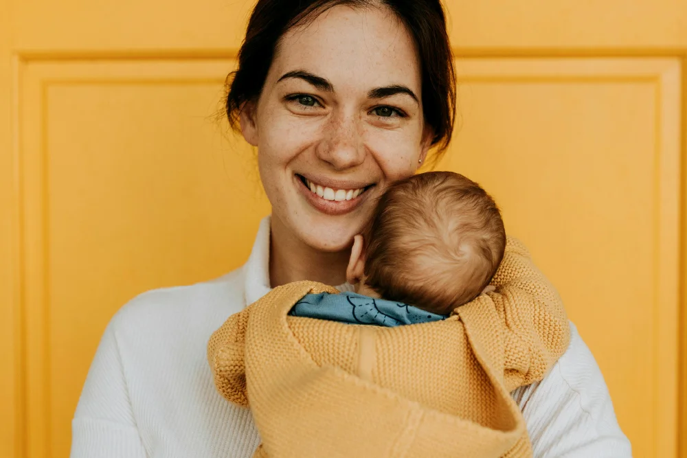 A woman with dark hair and freckles on her face is holding a newborn baby. She is looking at the camera and smiling