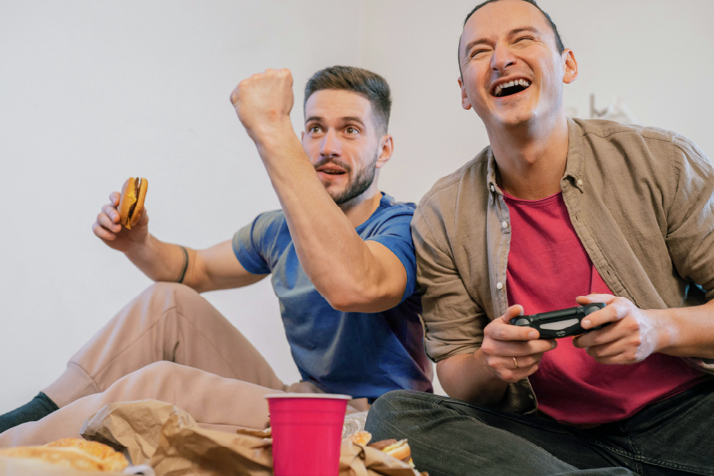 Two men enjoying video games together, one holding a controller and the other cheering with a fist in the air, both looking delighted.