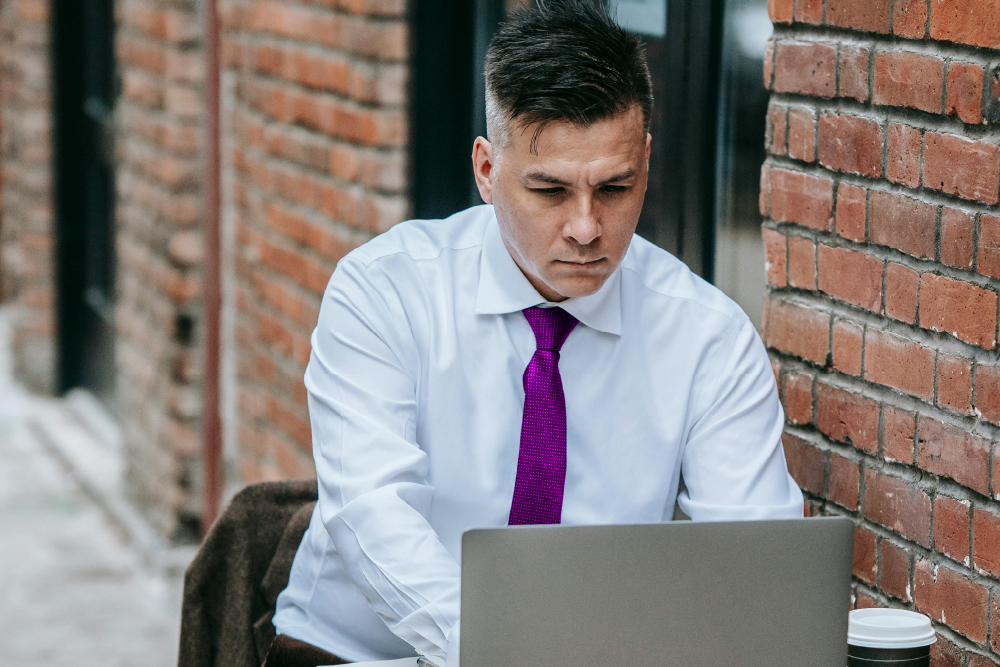A stressed looking man in a white shirt and purple tie working on a laptop outdoors, seated next to a brick wall.