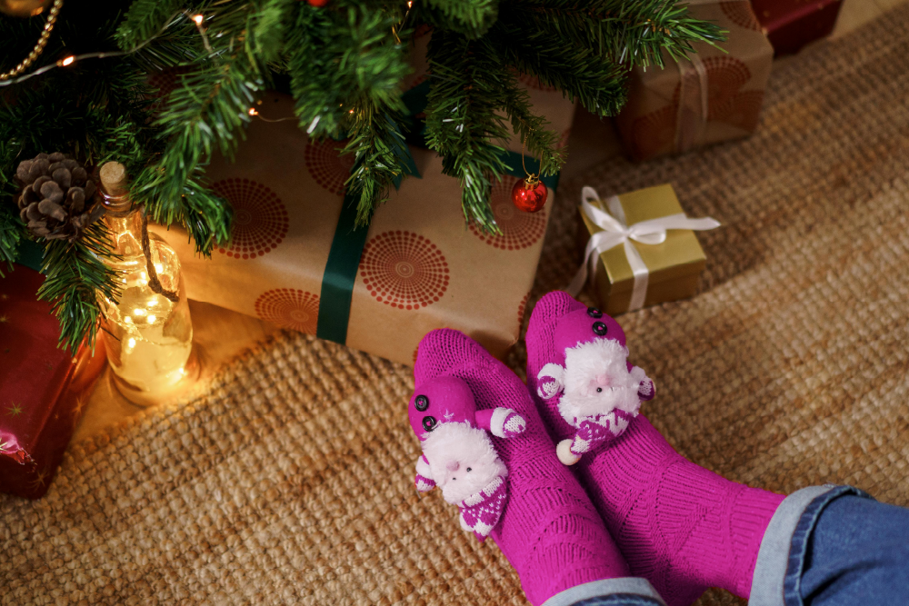 Pink socks with festive designs featuring plush toy characters, resting near a Christmas tree decorated with lights, presents, golden baubles, and pinecones on a woven rug