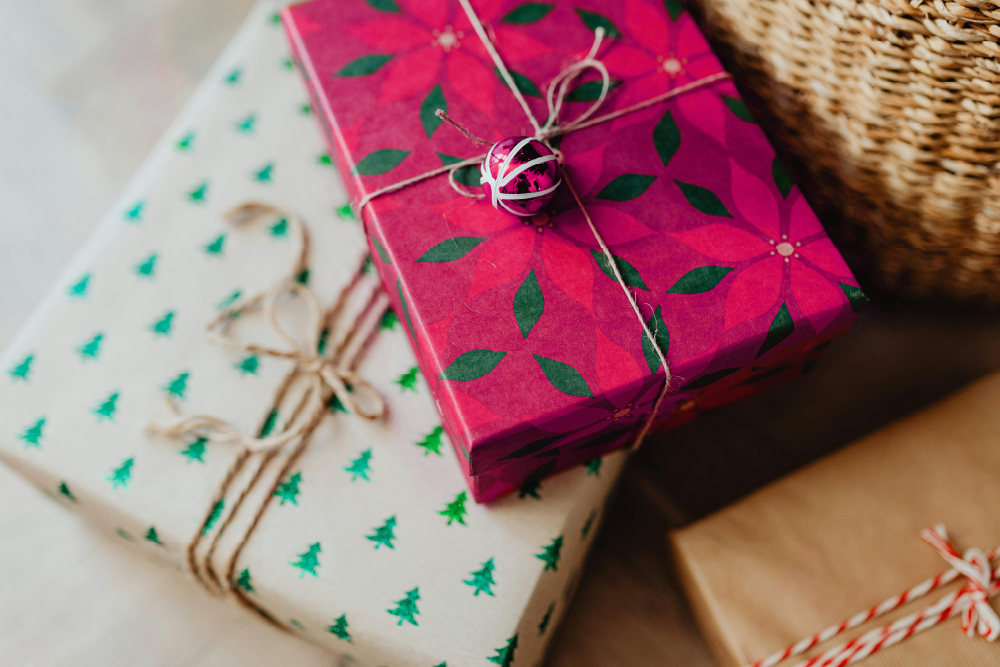 Festively wrapped gifts, including one with red and green poinsettia-patterned paper tied with twine and a small decorative bell, and another with white wrapping featuring green Christmas trees, placed near a woven basket