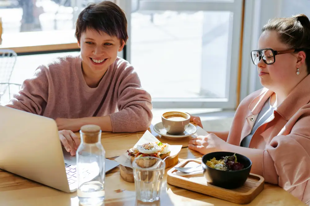 Two people are sitting at a wooden table, one using a laptop and is wearing a pink sweater while the other is wearing a pink jacket with white shirt and enjoying a coffee, surrounded by food and a water bottle.