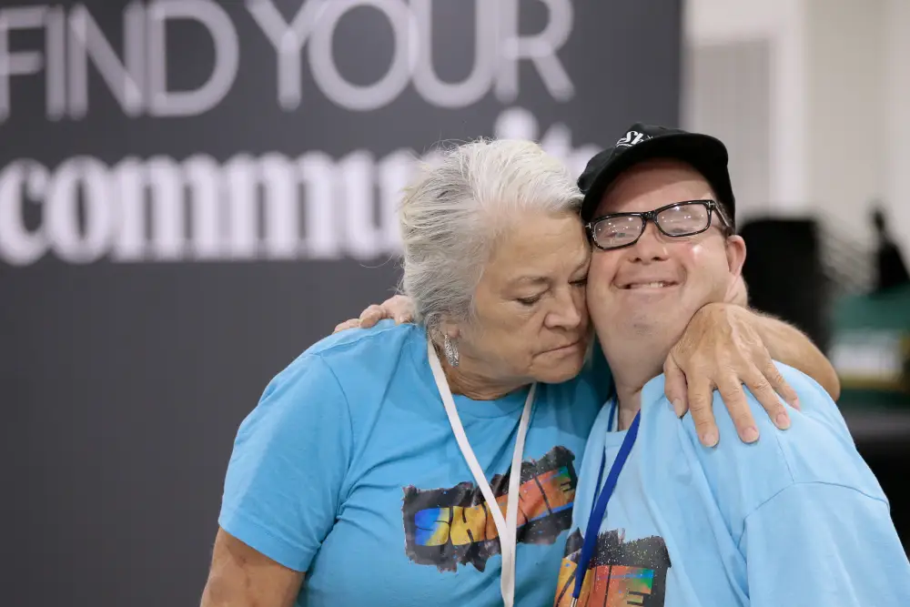 Two people in matching blue t-shirts share a joyful moment and hugging, standing in front of a backdrop that reads Find Your Community.