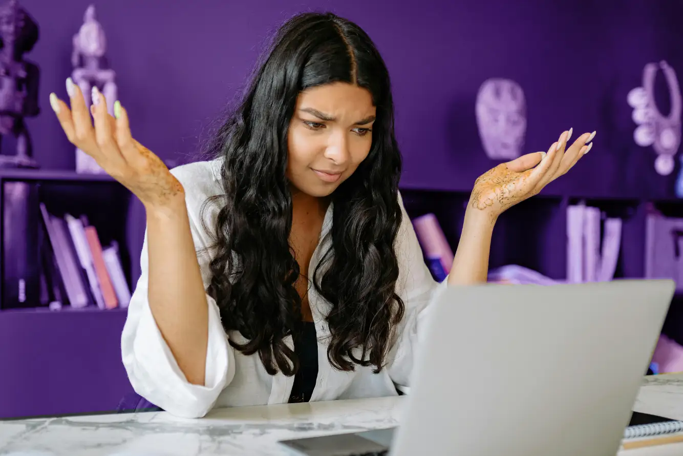 Woman with long dark hair and henna on her hands looking confused at a laptop, raising her hands in frustration.