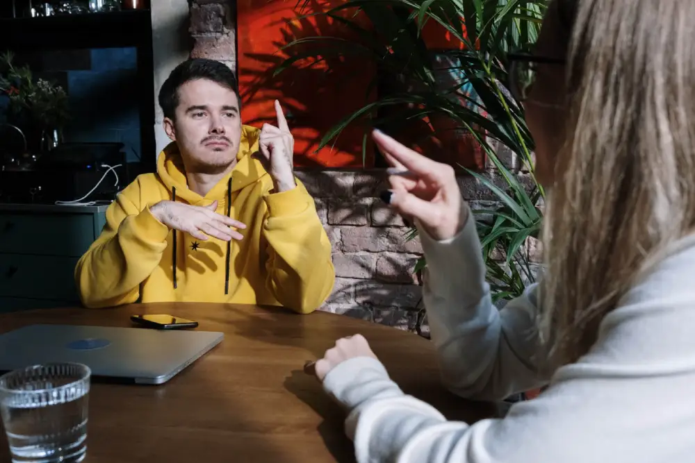 Two people using sign language at a wooden table, one wearing a yellow hoodie and the other in a light-colored sweater