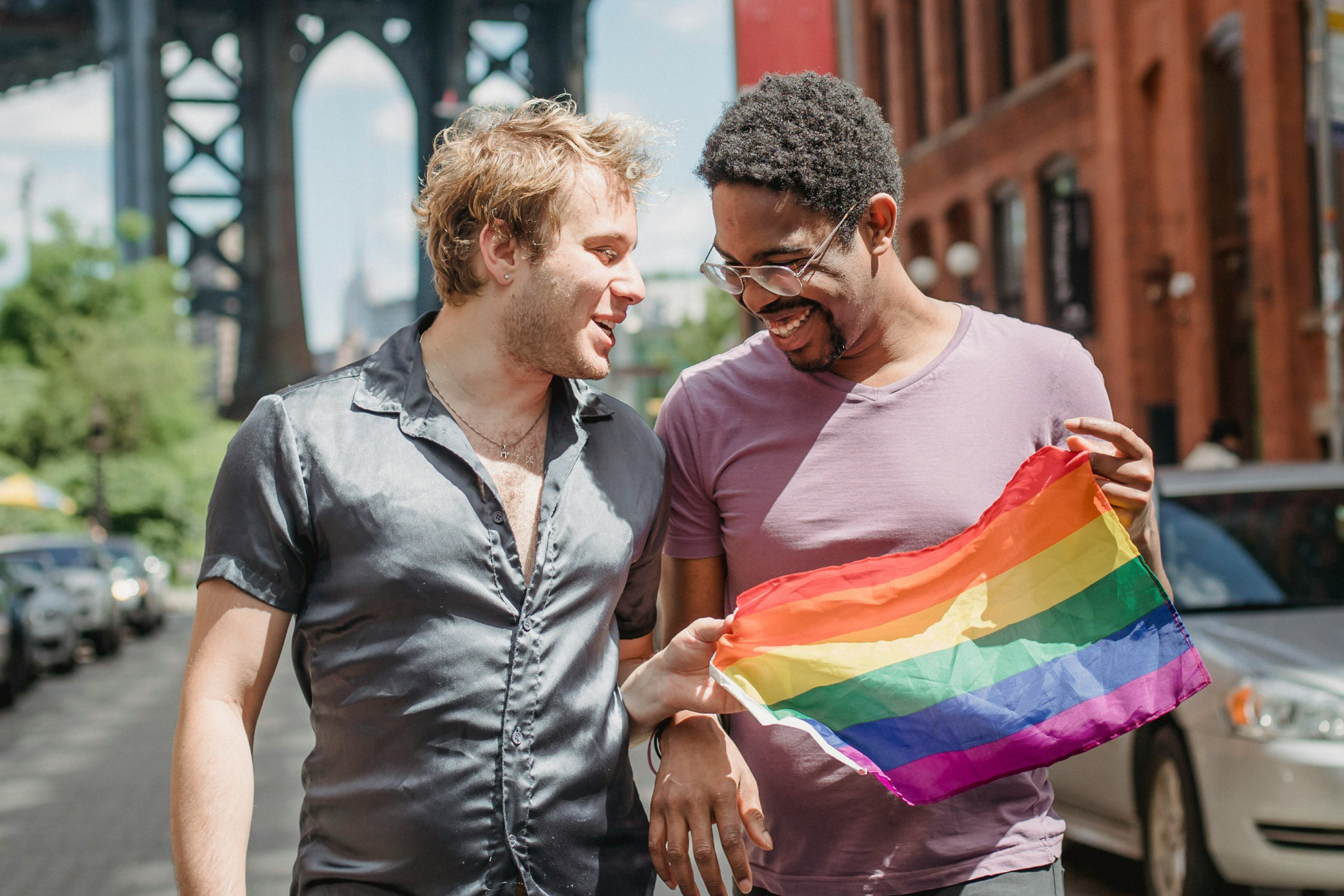 Two people smiling in conversation in a city street. Together, they are holding a Rainbow Pride flag.