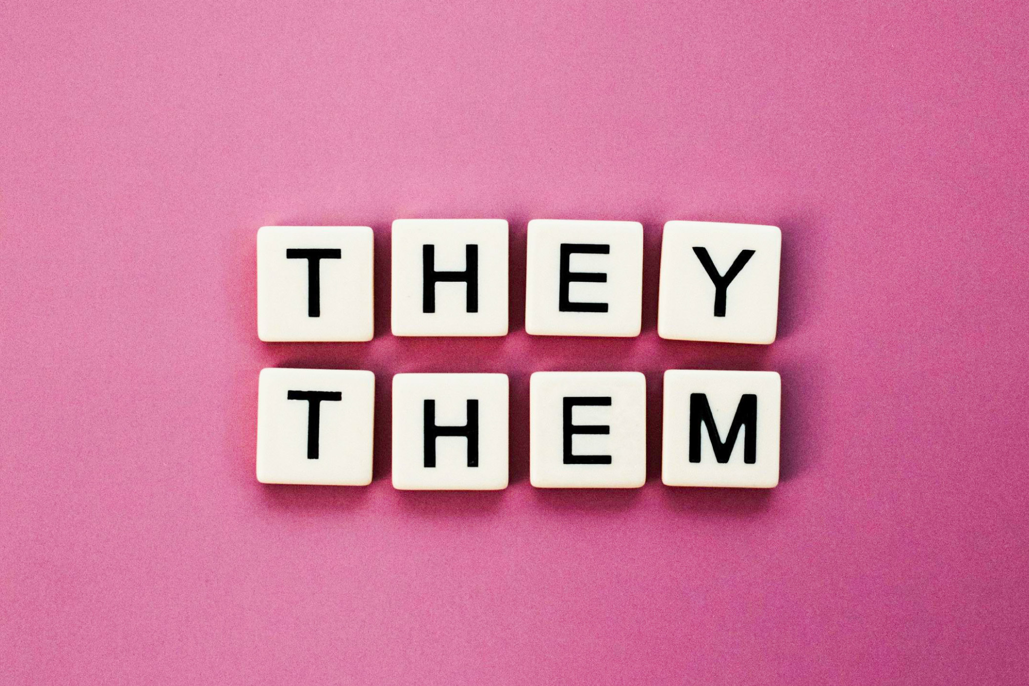 Eight Scrabble tiles arranged to spell the words ‘they them’ against a pink background.