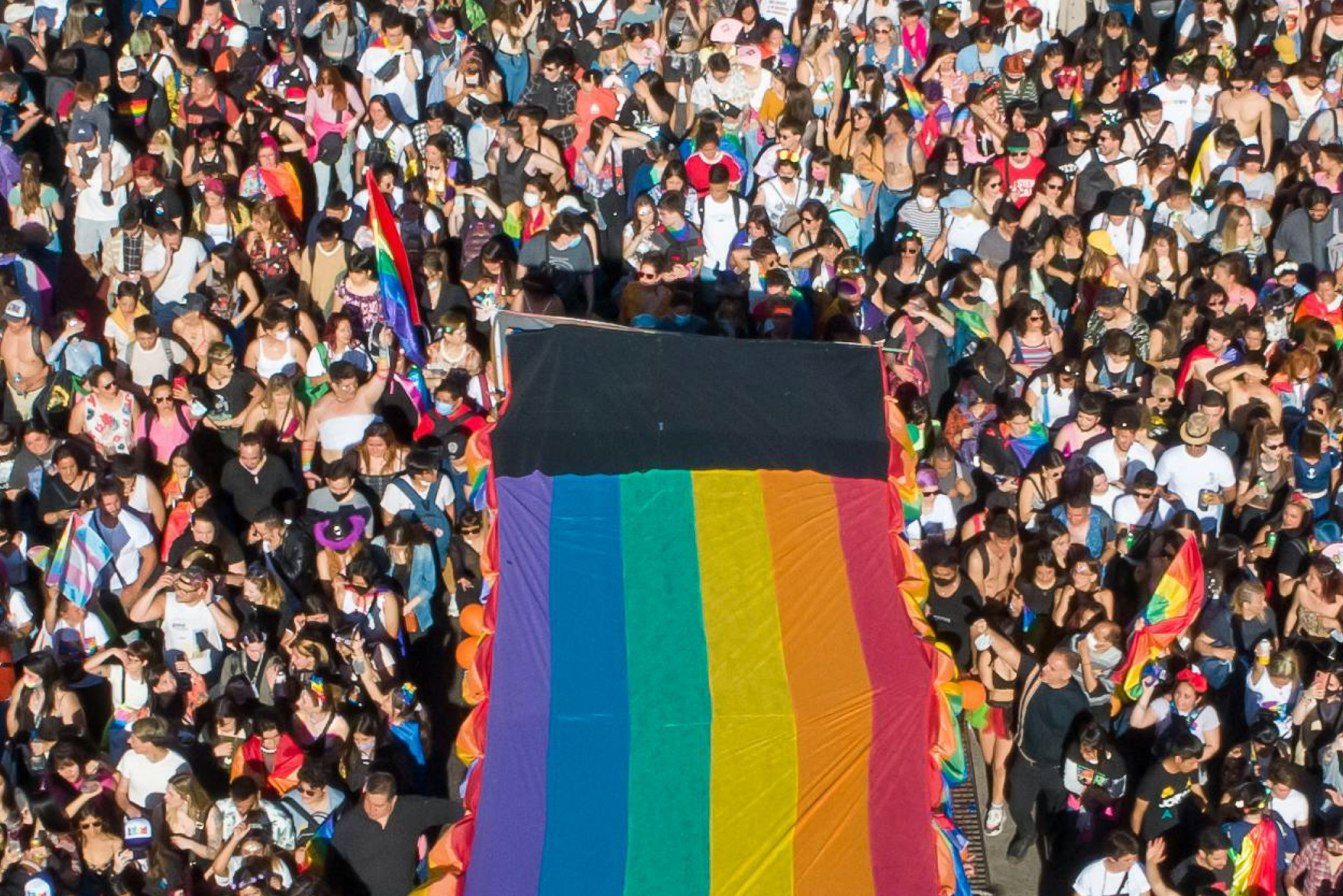 A crowd of people gathered for a Pride parade, with a large Rainbow Pride flag unfurled in the centre.