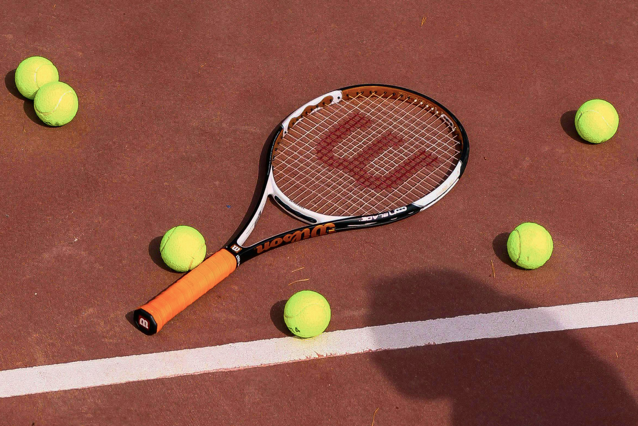 An orange, yellow and black tennis racket and six yellow tennis balls laid out against the burgundy surface of a tennis court with a white painted stripe. The shadow of a person’s head is in the bottom right corner