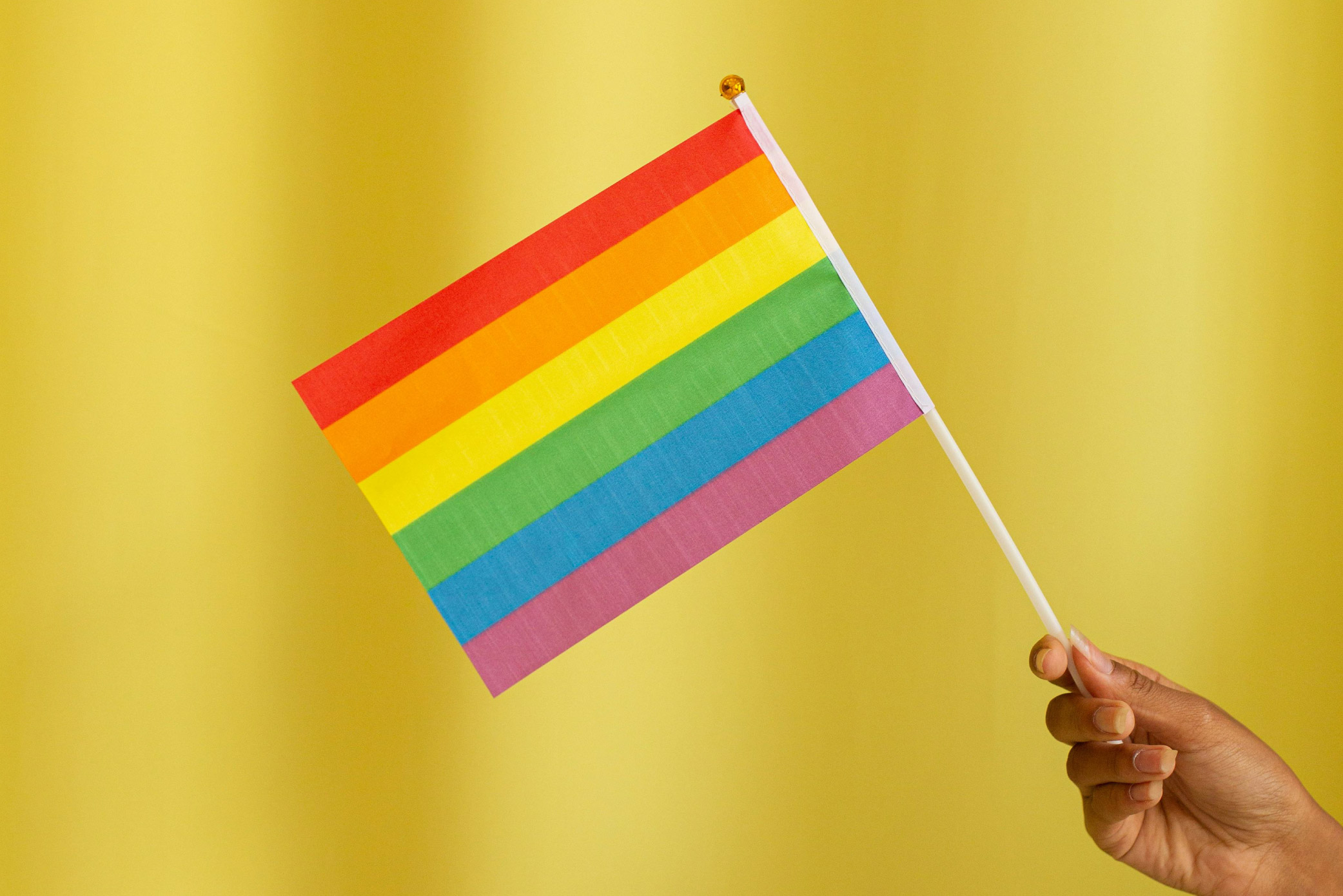 A hand holding a miniature Rainbow Pride flag against a gold background.
