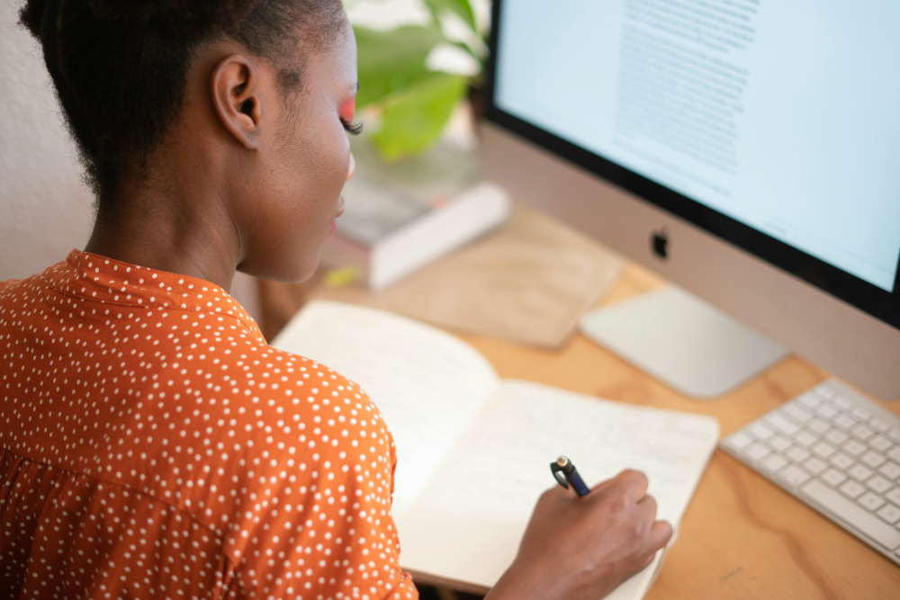 A person is sitting at a desk in front of a computer screen, using a pen to write in a notebook