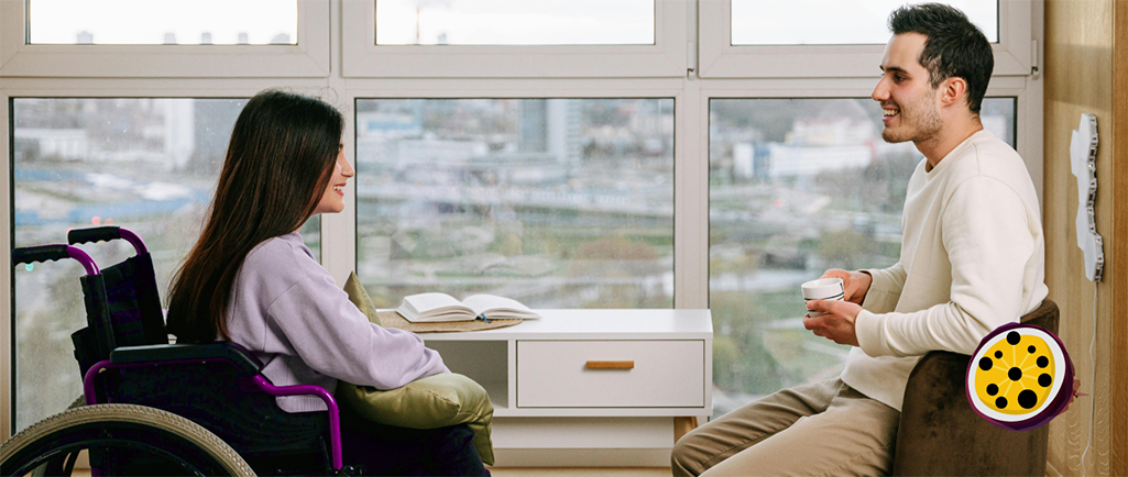 Two colleagues are conversing, seated in an office in front of a window. One is a wheelchair user wearing a pale blue long-sleeved top with long, dark hair. The other is holding a coffee cup, wearing a white long-sleeved top and stone coloured trousers, with short dark hair.