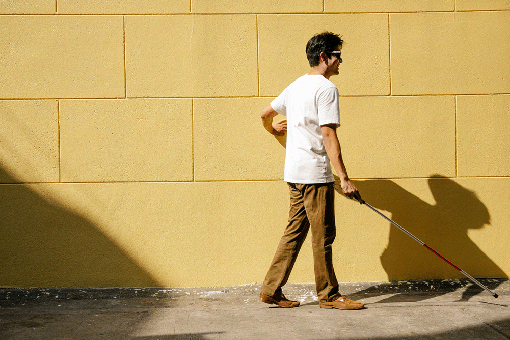 A blind person is walking on a grey, concrete pavement using an assistive cane. Their hand is placed against the beige brick wall beside them. The sunlight casts their shadow on the wall and pavement ahead of them.