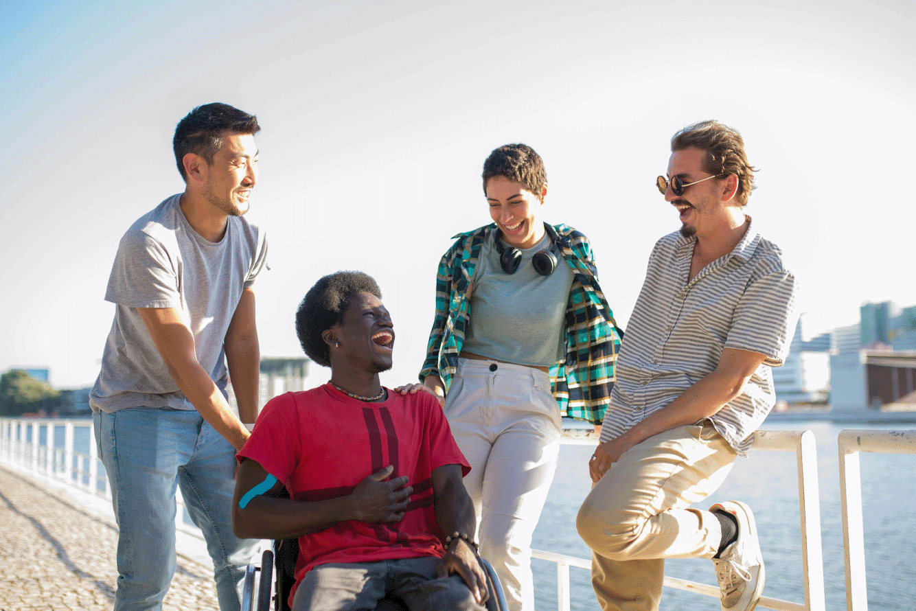 Four people conversing outside in the sunshine, with a river and blue skies behind them in the background. They are all dressed casually and have short, dark hair. Two people in the group are leaning against a white fence and laughing. The other two people in the group are facing them. One is a wheelchair user, laughing with their hand on their belly, the other is smiling with their hand on the wheelchair.