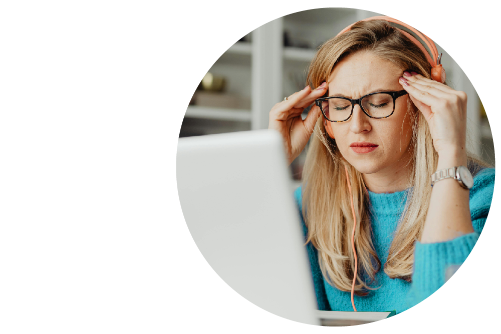 Person wearing headphones and glasses at laptop, eyes closed with both hands pressed to temples, appearing stressed