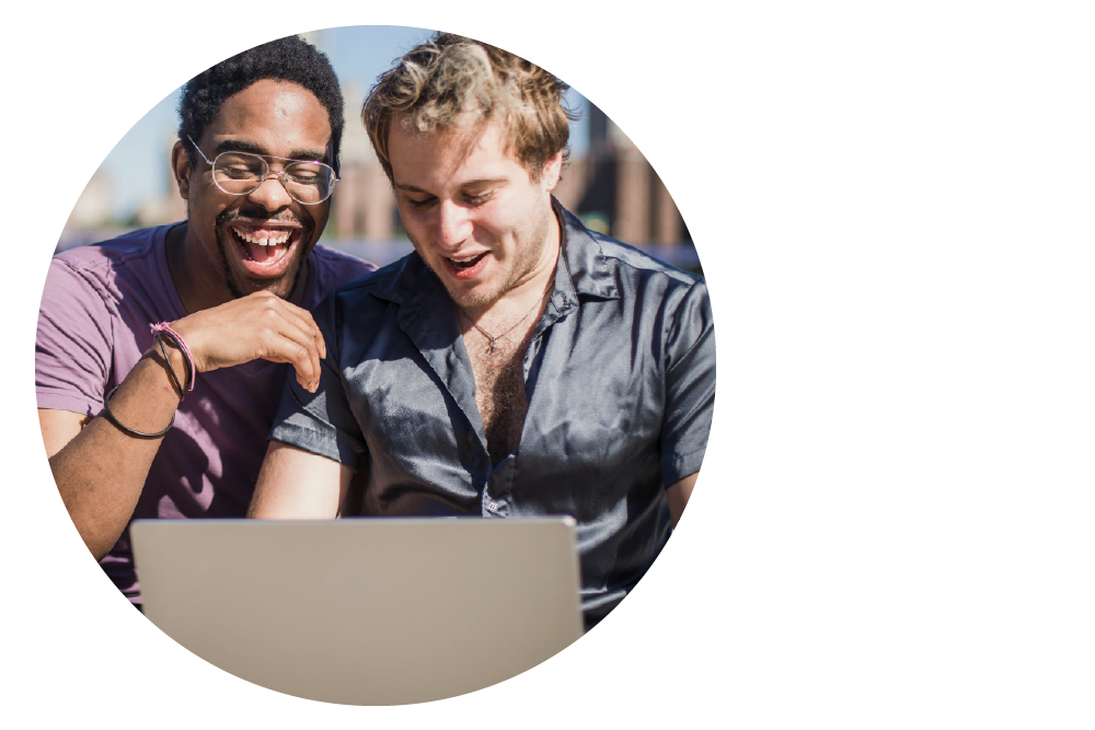 two people sitting close together outdoors and laughing animatedly while looking down at a laptop computer.