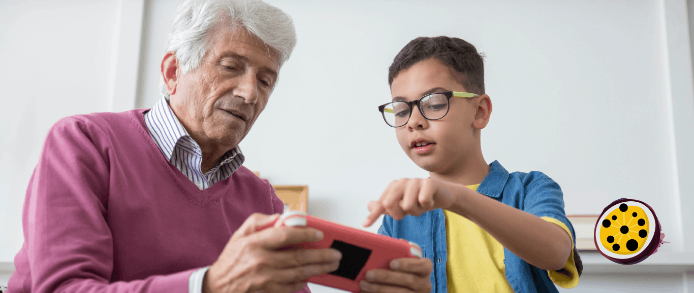 An adult and a child are using a pink and black handheld device. The adult is holding the device and looking down at the screen. They have short, grey hair. They are pointing to the device screen.