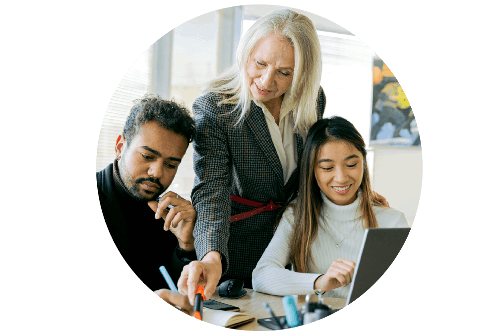 Senior woman mentoring two coworkers at a desk, pointing at notes while one uses a laptop and the other writes.