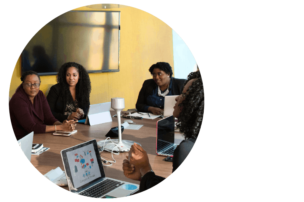 four Black people sitting around a table. They are dressed in business attire with laptop computers and tablet devices on the table, and interested expressions as if engaged in conversation.