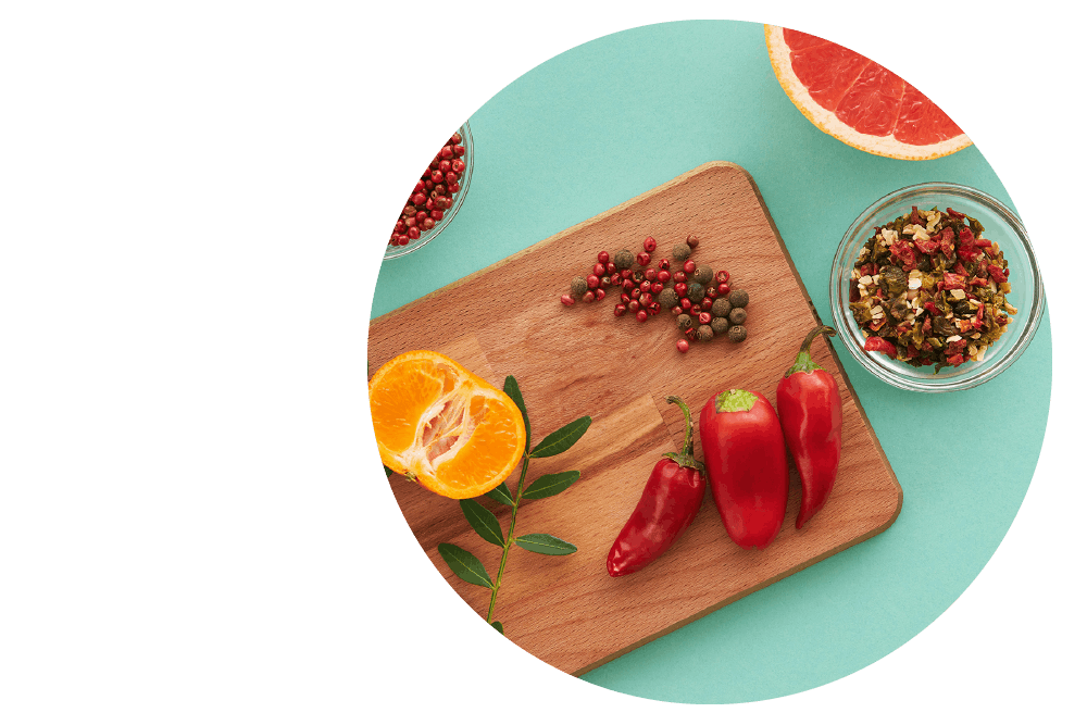 Red chili peppers, orange slices, and spices on a wooden board over teal background.