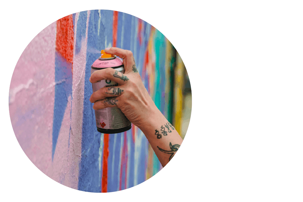 a tattooed wrist and hand using a can of spray paint on a wall decorated with brightly coloured shapes.