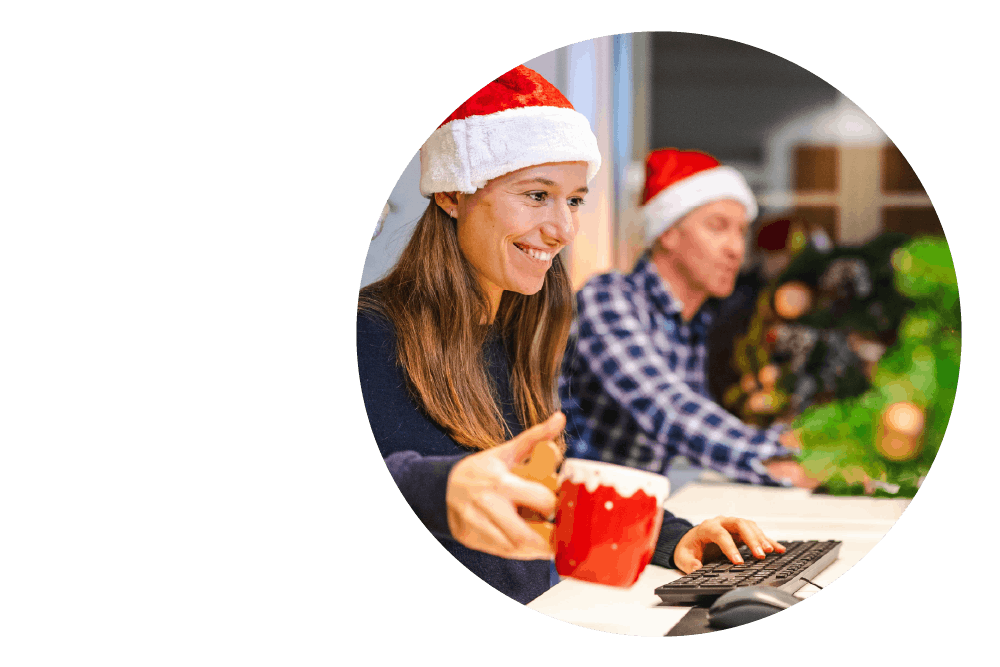 a smiling person wearing a Santa hat and sitting at a desk. They are holding a festive red mug in one hand and using a computer keyboard with the other.