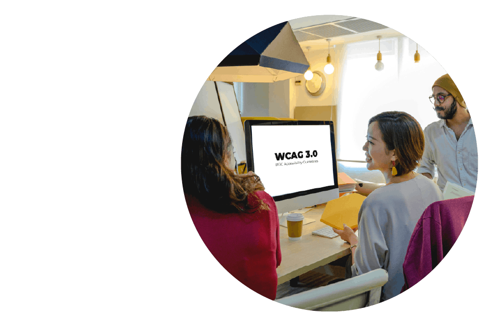 three people gathered around a desk with a computer. The first person has their back to the camera, the next is facing them and smiling, and the third person is looking at the computer. Text on the computer’s screen reads: WCAG 3.0