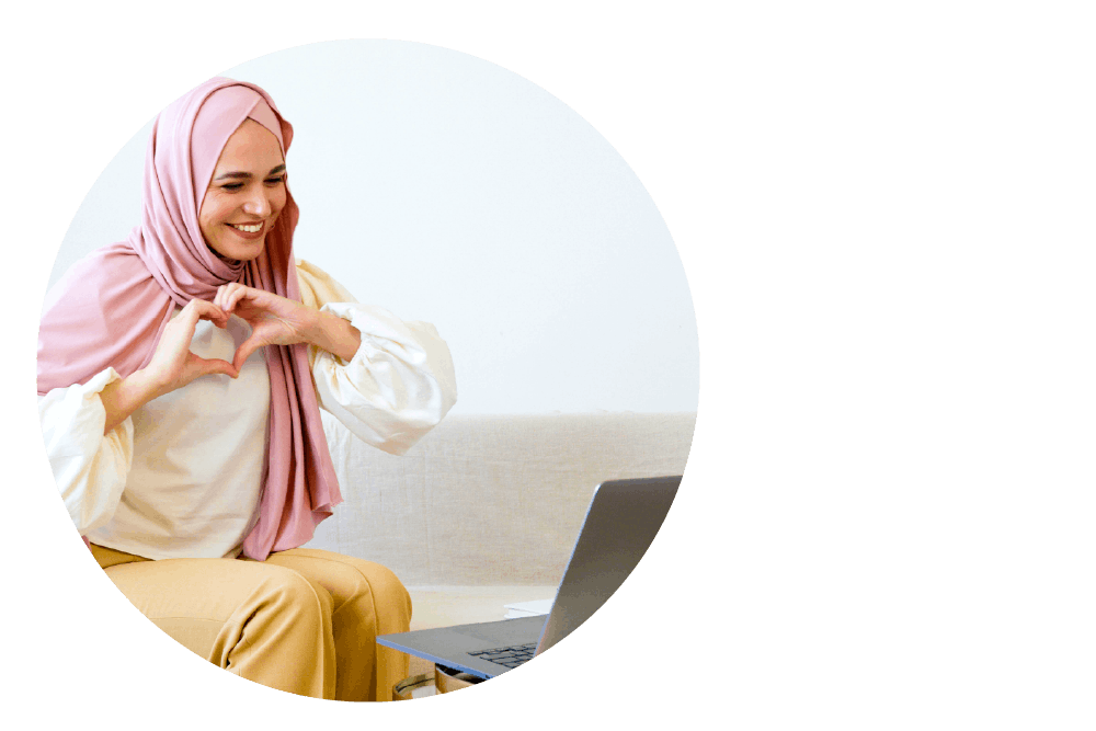 a person wearing a headscarf and looking down at a laptop computer. They are smiling and using their hands to form a heart shape against their chest.