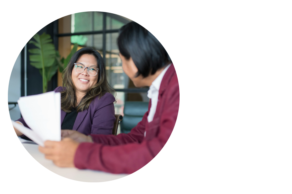 Aligned to the left, a circle frames two women sitting at a desk holding paper documents, looking at each other and smiling
