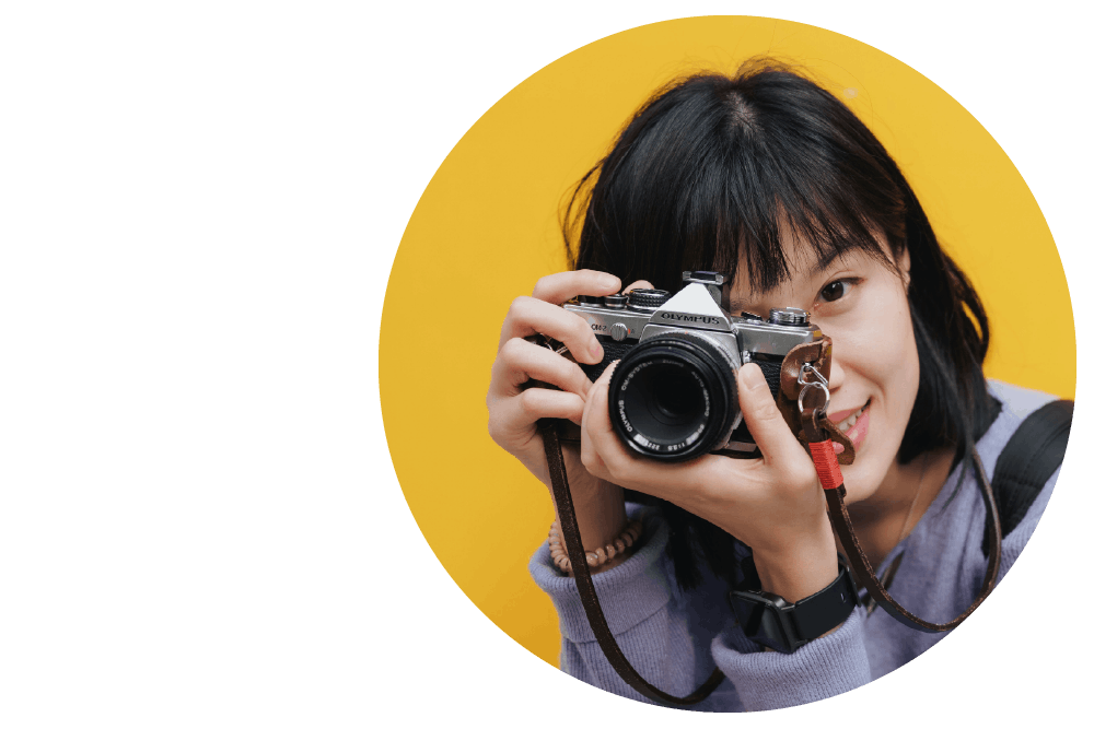 a smiling person looking through the lens of a film camera held up close to their face against a yellow background