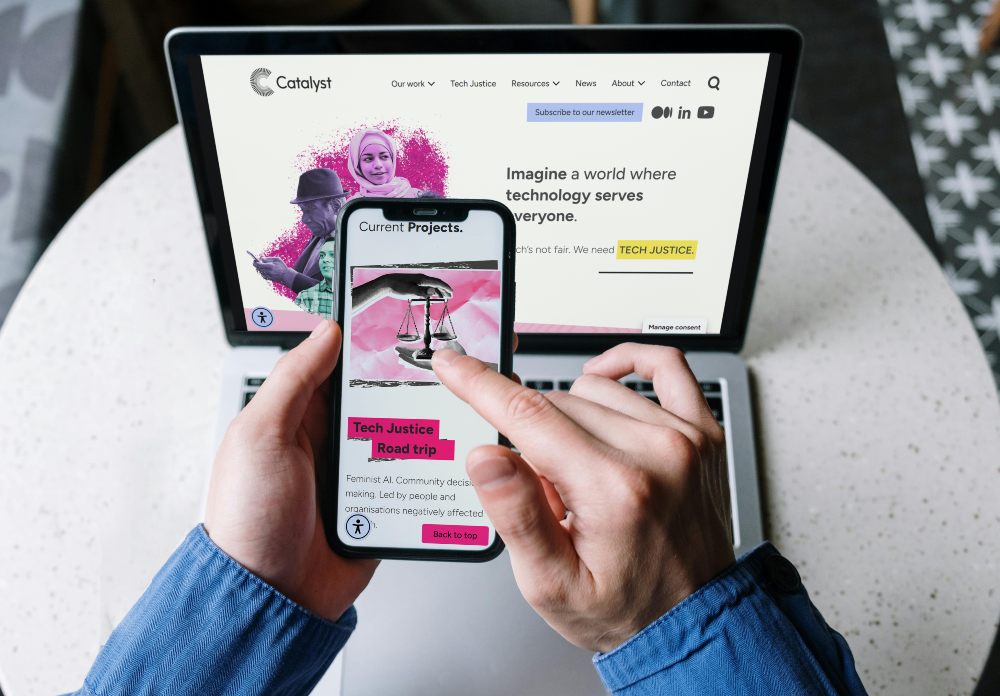 Person using a smartphone in front of a laptop, viewing the Catalyst website and a Tech Justice project page. The laptop displays Catalyst’s homepage with illustrations and text about technology and social justice, while the smartphone shows project details. Both devices are placed on a round table.