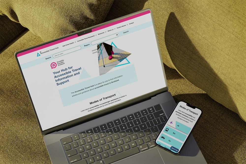 A person holds a smartphone displaying an accessible travel information website while sitting in front of an open laptop showing the same site. The phone screen lists transportation options including air, bus, and car, and a glass of water sits beside the laptop