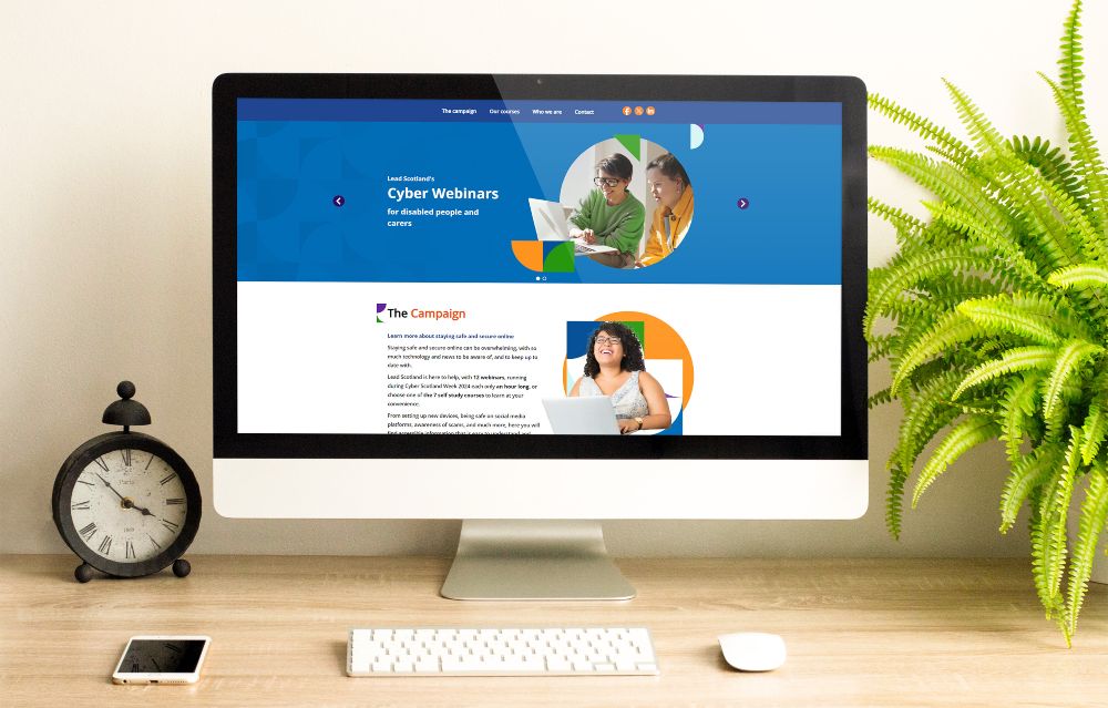 Desktop computer displaying a blue cyber webinars website on a wooden desk with a keyboard, mouse, smartphone, vintage clock, and green fern plant