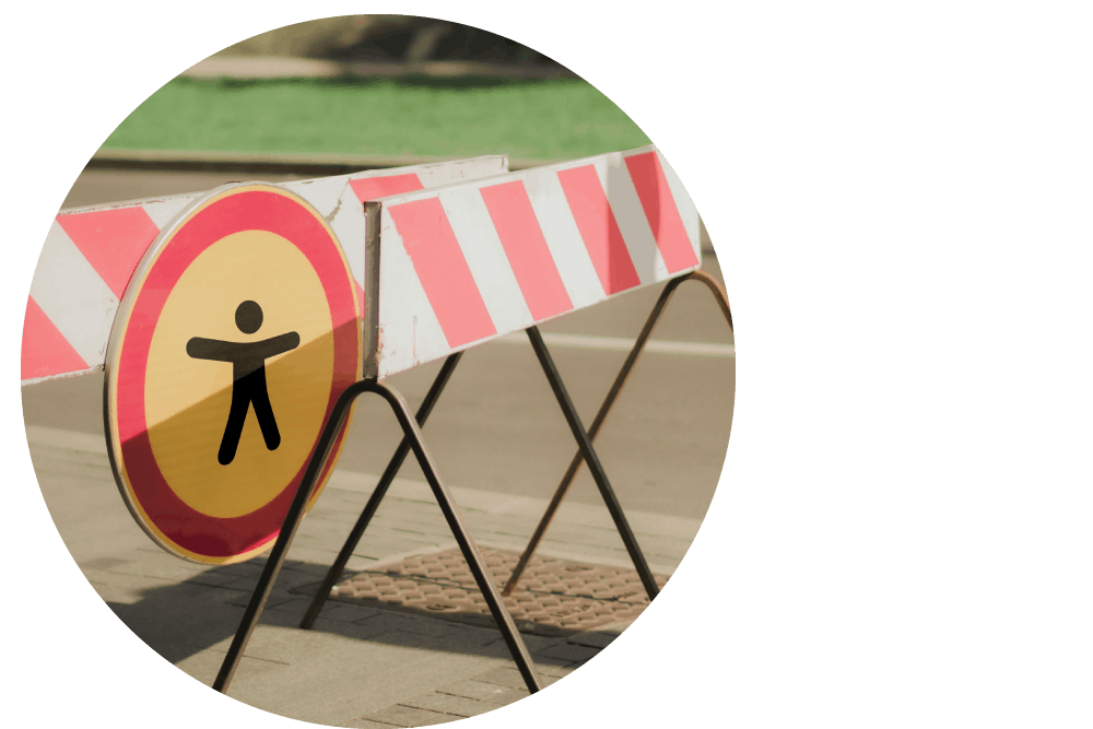 Red and white striped barriers blocking a pavement outdoors. A red and yellow circular warning sign displays a black accessibility icon.