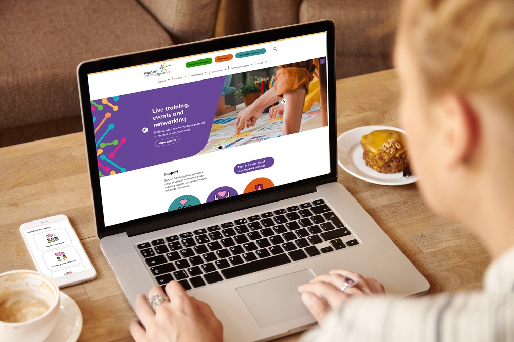 Person using a laptop on a wooden table, viewing Support Cambridgeshire website with colorful banners and navigation menus. A smartphone displaying related content sits beside the laptop, with a cup of coffee and a slice of cake nearby.