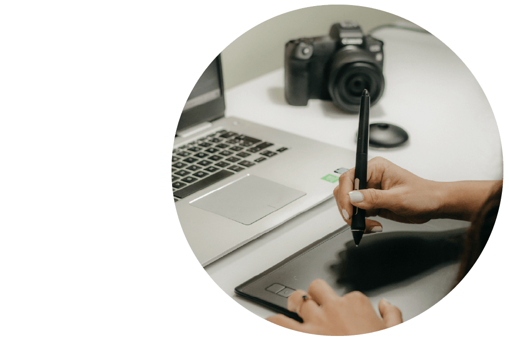 Hands using a stylus with an open laptop computer and an SLR camera on a surface in the background.
