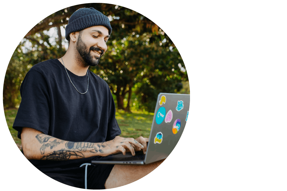 A person sitting outdoors using a laptop computer, looking down at the screen and smiling.