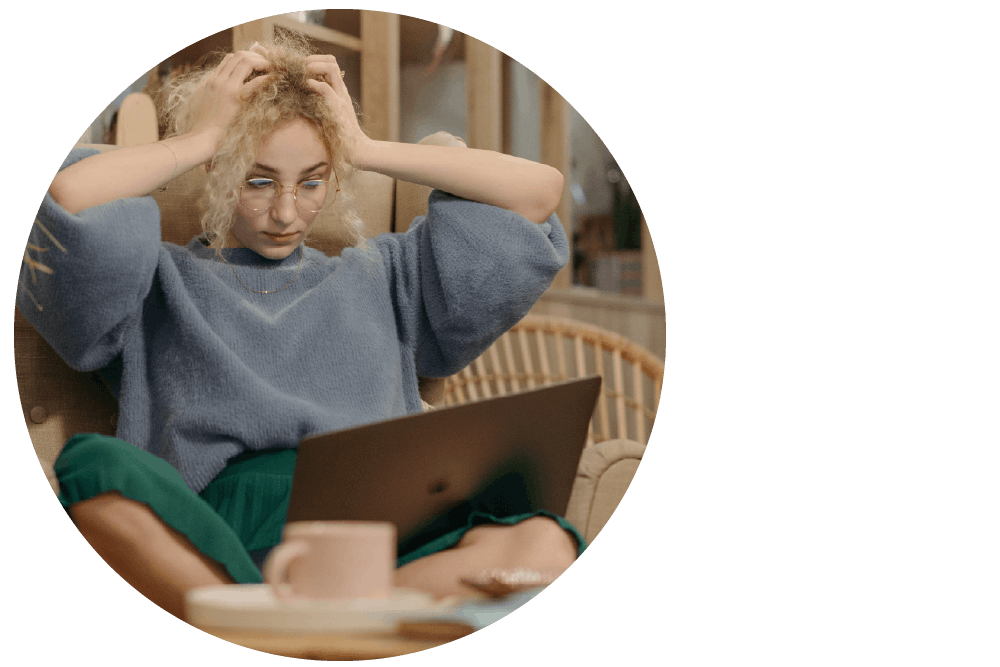 A person sitting cross-legged with a laptop computer in their lap, looking down at the screen with both hands raised to their head and a frustrated expression.