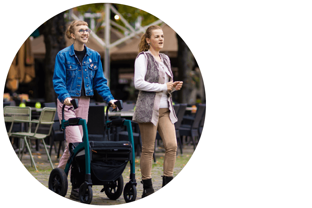 Two people walking side by side outdoors and smiling widely, one of whom is using a mobility aid.